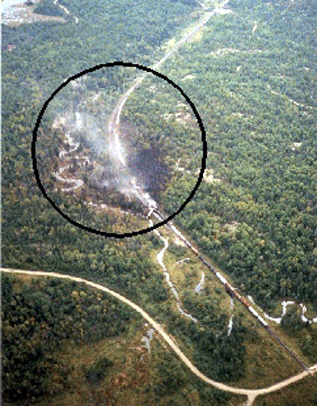 Figure 3 - Aerial photograph of derailment site from the northwest. The circle denotes blackened area from fire damage. Note the public crossing at the bottom right. Note the direction of smoke and fumes, and the location of Little Key River adjacent to the right-of-way.